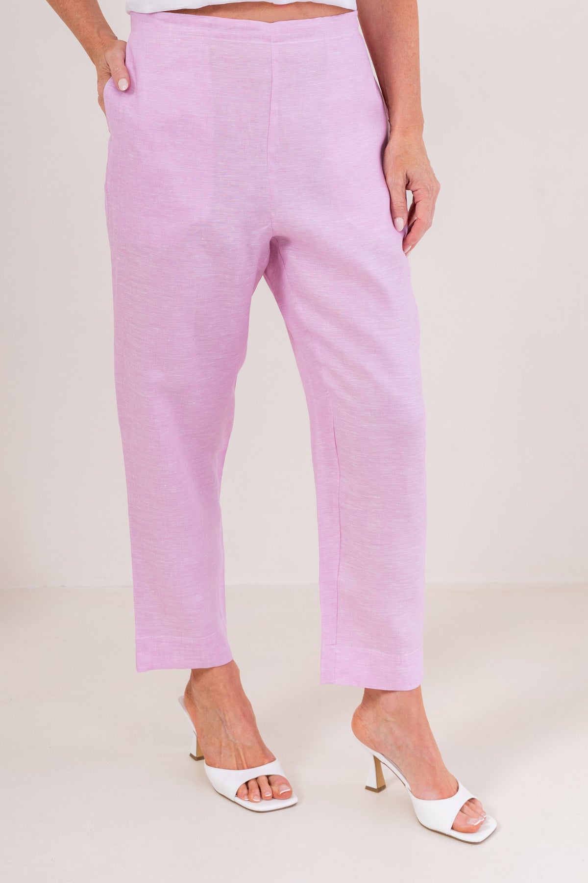 A person wearing light pink linen  See Saw 7/8 pants with a relaxed straight-leg fit, paired with white heeled slide sandals, photographed against a plain light beige studio background.