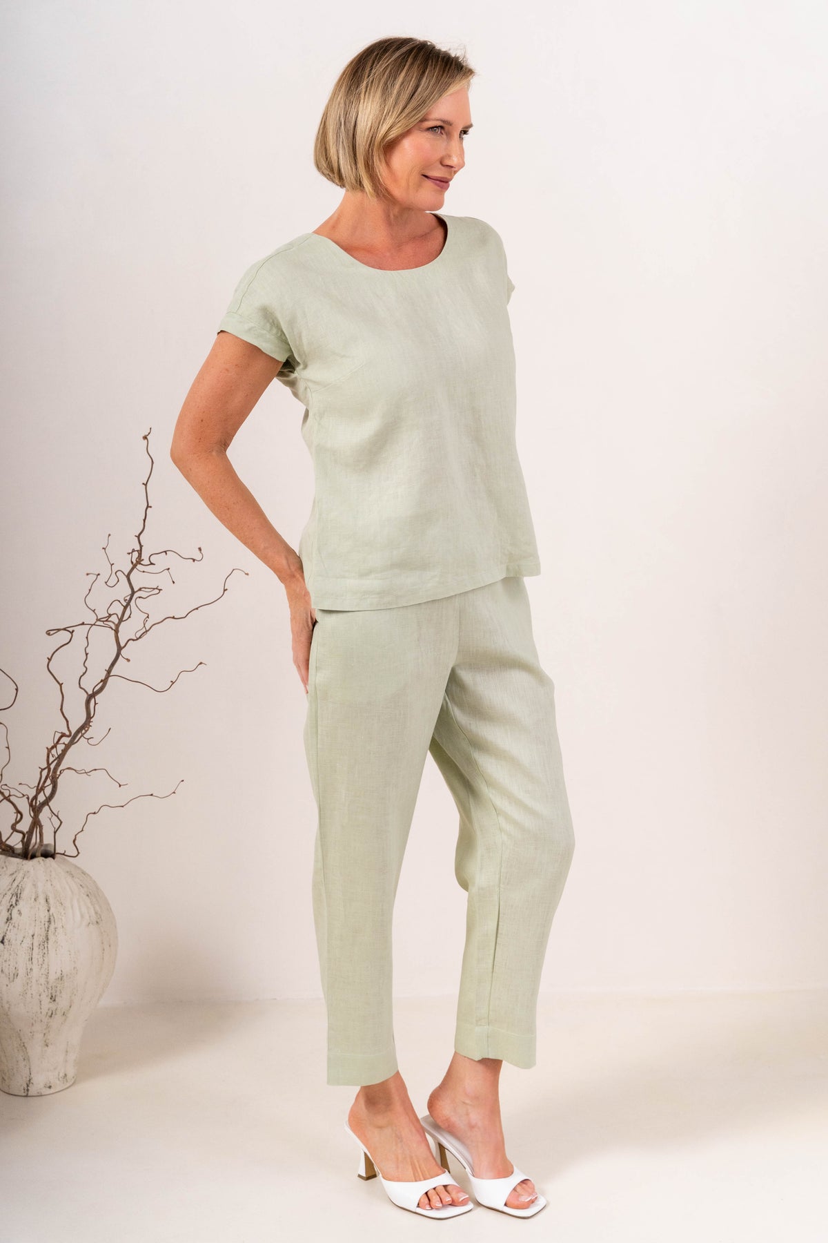 A person wearing light sage linen  See Saw 7/8 pants with a relaxed straight-leg fit, paired with white heeled slide sandals, photographed against a plain light beige studio background.