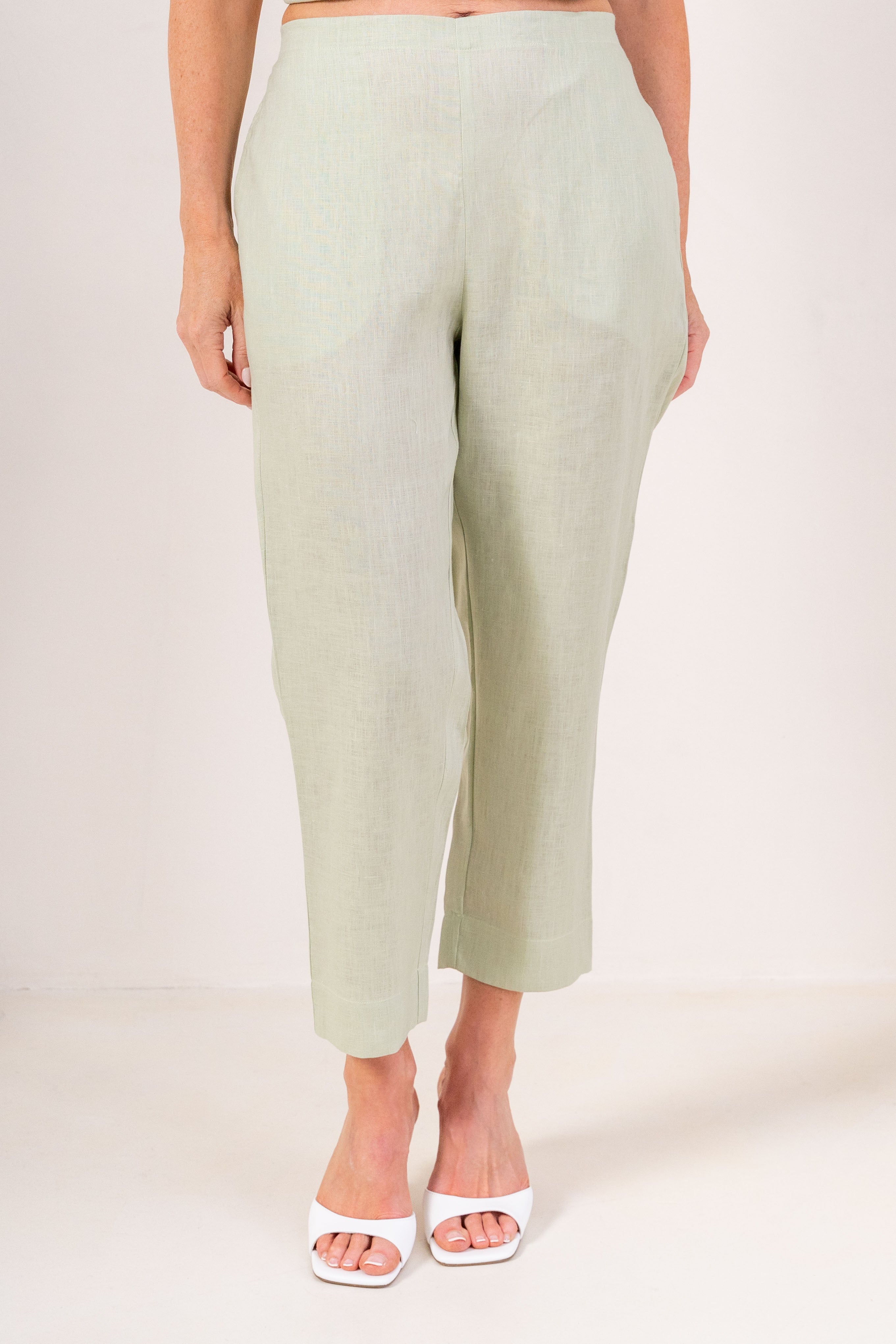 A person wearing light sage linen  See Saw 7/8 pants with a relaxed straight-leg fit, paired with white heeled slide sandals, photographed against a plain light beige studio background.