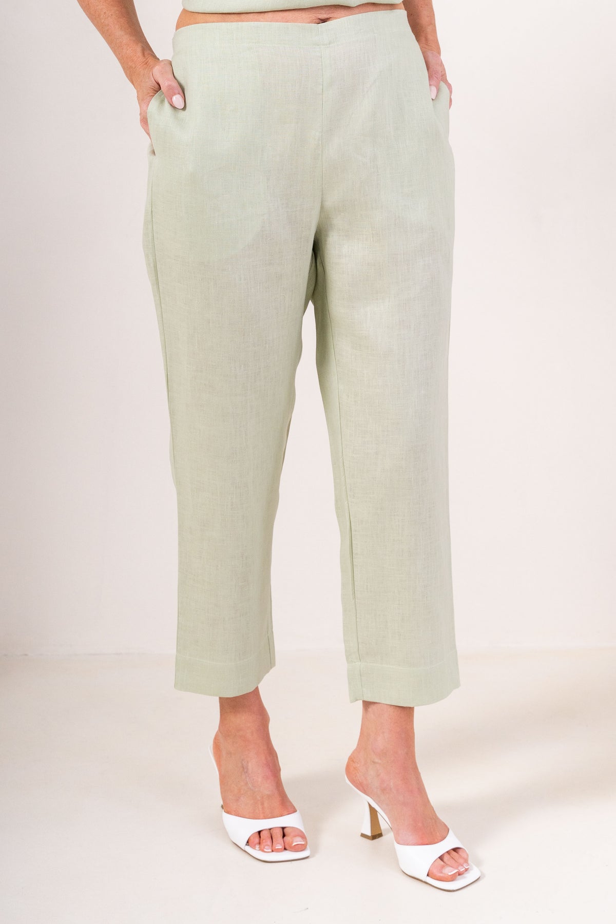 A person wearing light sage linen  See Saw 7/8 pants with a relaxed straight-leg fit, paired with white heeled slide sandals, photographed against a plain light beige studio background.
