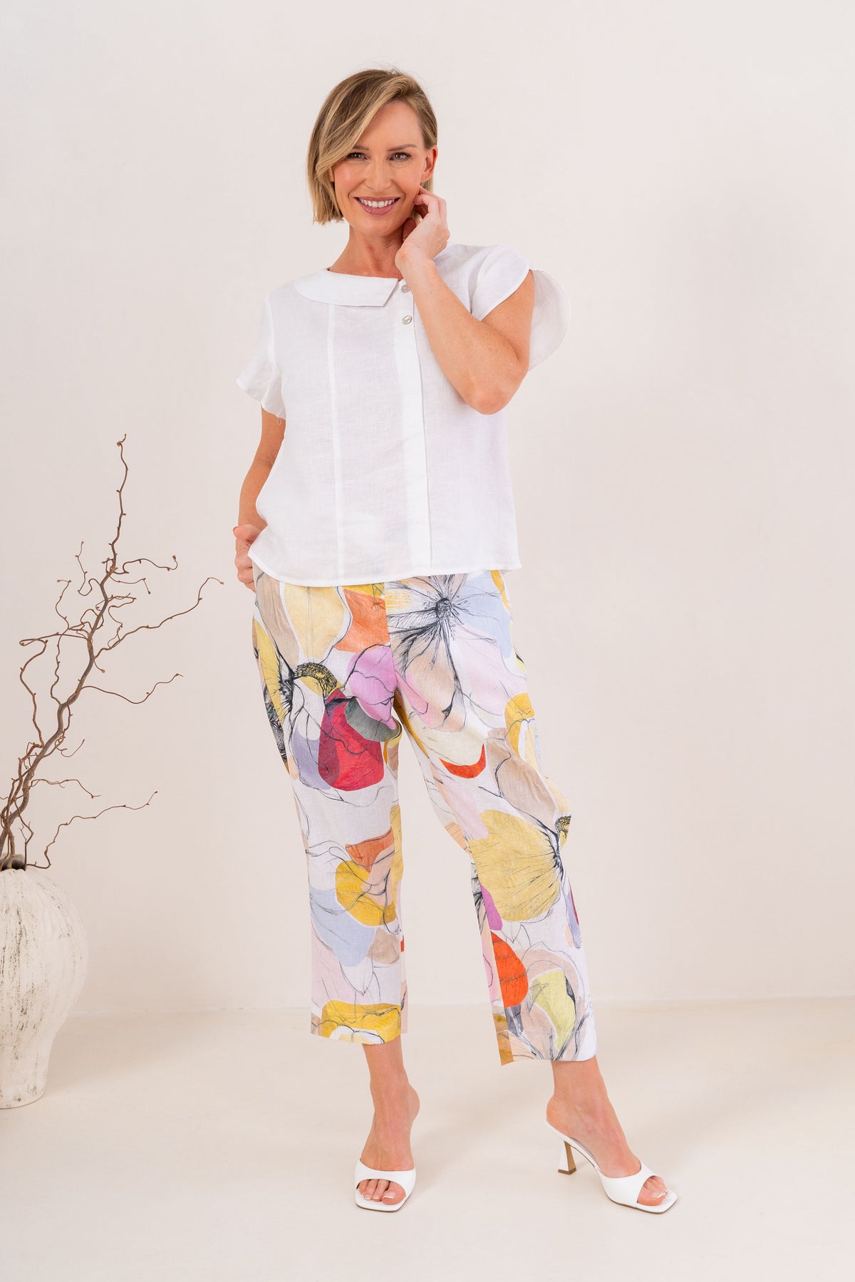 A person wearing See Saw linen cropped pants with a large abstract Petal Drift print in yellow, pink, orange, purple, and blue on a light base, paired with white heeled sandals against a plain beige studio background.