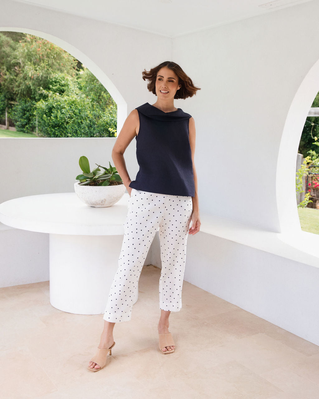 A woman modeling a sleeveless linen See Saw dark navy top and white pants with small black polka dots, standing in a bright, modern white room with a curved table and greenery visible through an arched window.