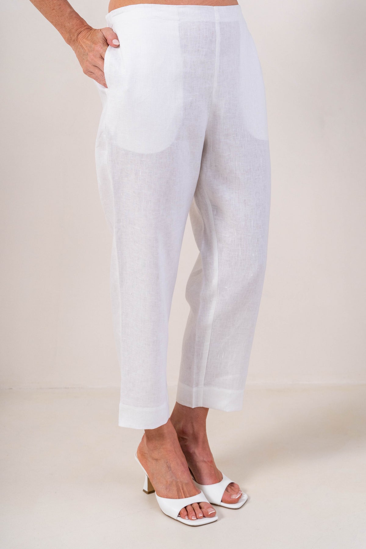 A person wearing white linen  See Saw 7/8 pants with a relaxed straight-leg fit, paired with white heeled slide sandals, photographed against a plain light beige studio background.