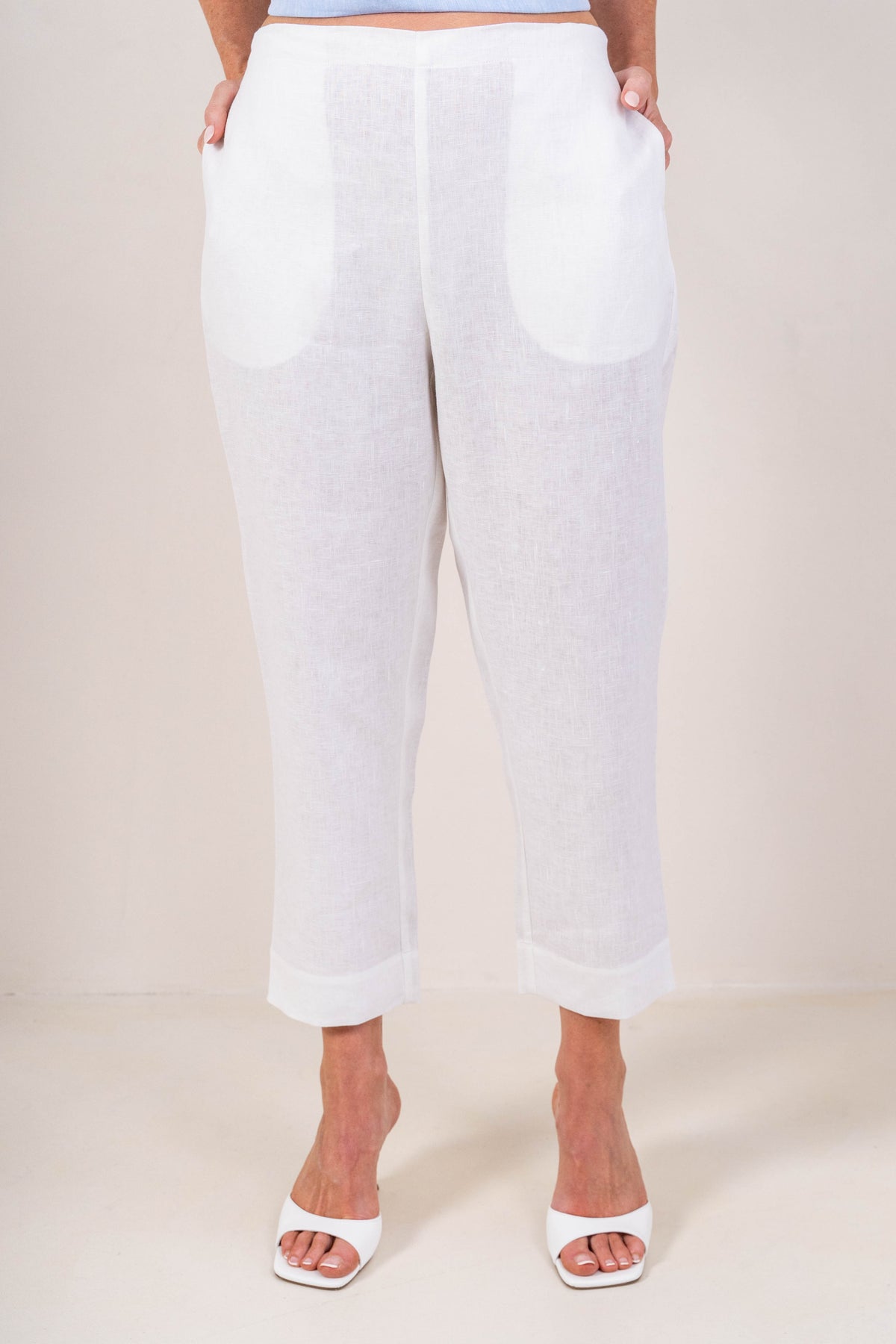 A person wearing white linen  See Saw 7/8 pants with a relaxed straight-leg fit, paired with white heeled slide sandals, photographed against a plain light beige studio background.