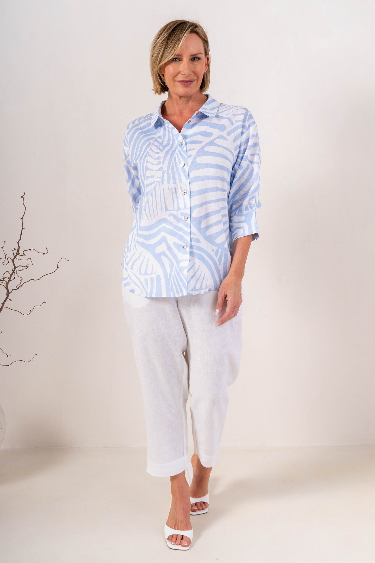 A person wearing white linen  See Saw 7/8 pants with a relaxed straight-leg fit, paired with white heeled slide sandals, photographed against a plain light beige studio background.