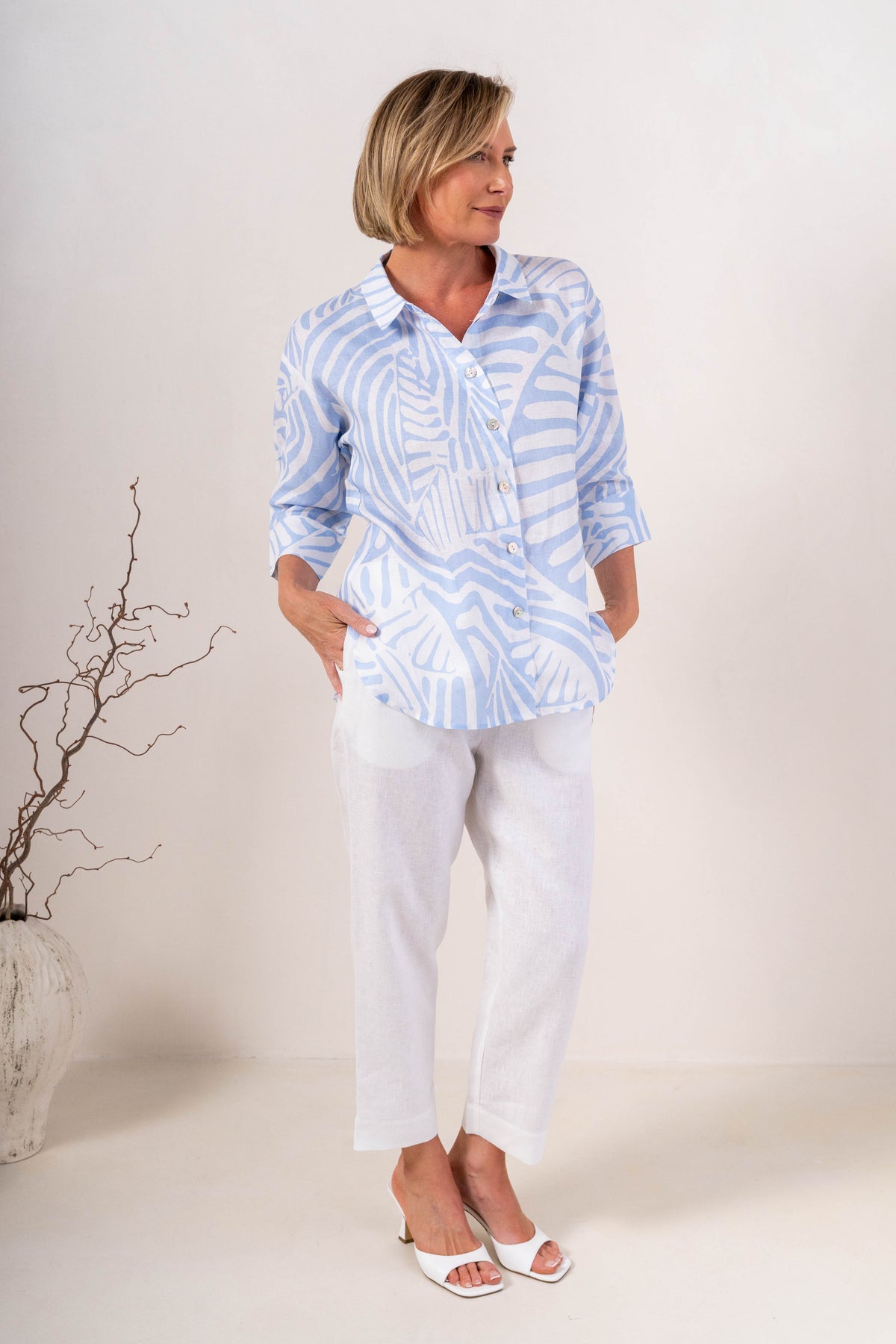 A person wearing white linen  See Saw 7/8 pants with a relaxed straight-leg fit, paired with white heeled slide sandals, photographed against a plain light beige studio background.