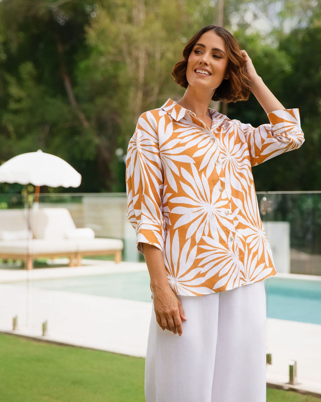 A woman wearing a Linen See Saw Summer Days print button-up shirt and white pants, standing outdoors in front of a swimming pool with green trees in the background.