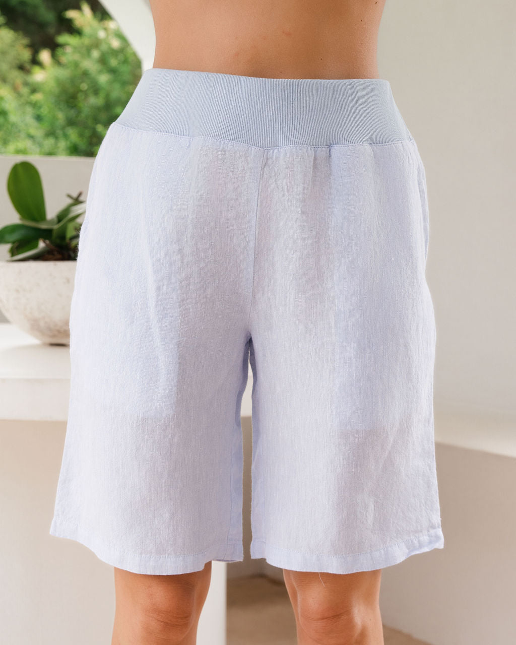 Light blue linen-blend See Saw linen shorts with a contrasting light blue elastic waistband, shown on a model from the waist down in a bright indoor setting.