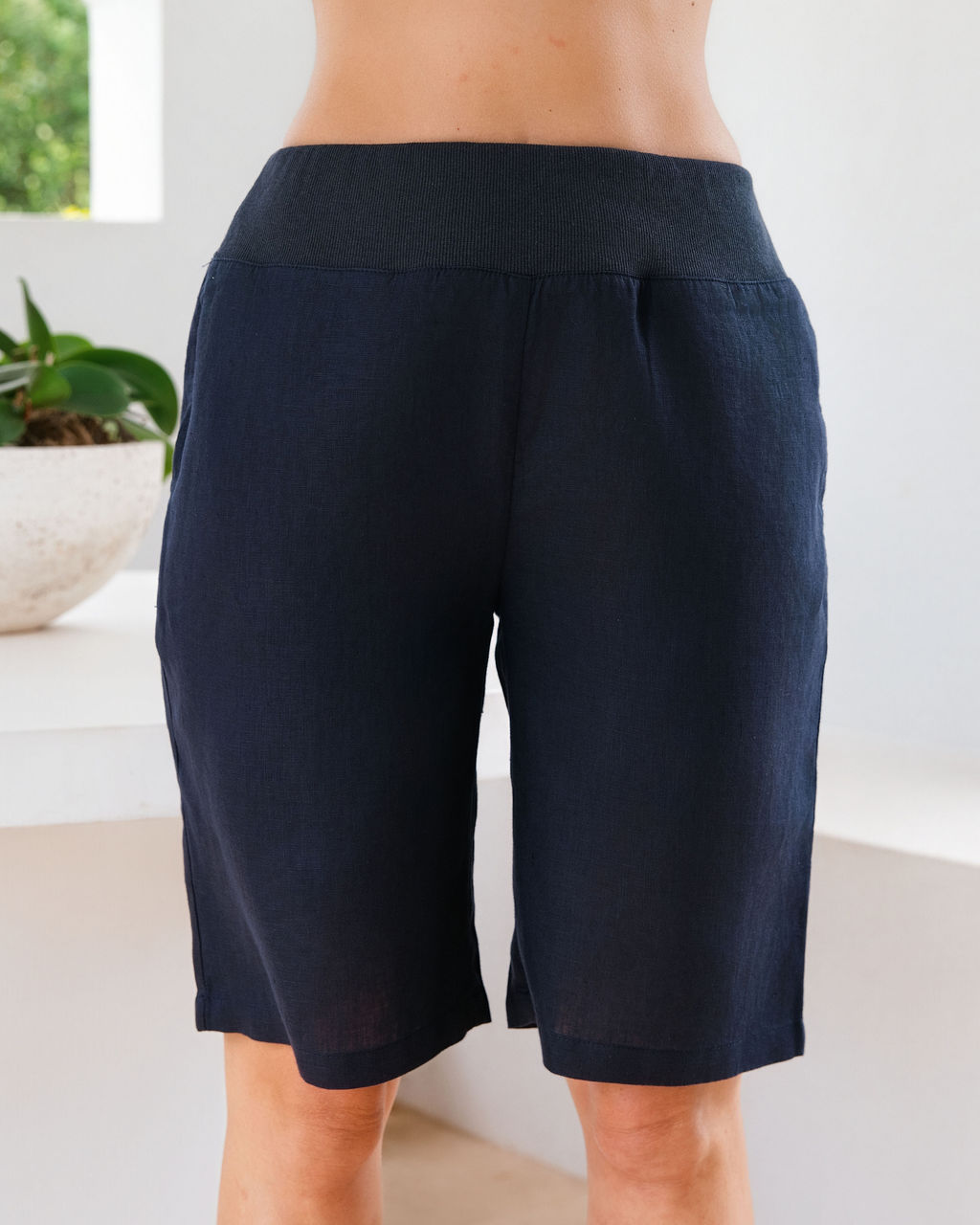 Navy linen See Saw linen shorts with a contrasting light blue elastic waistband, shown on a model from the waist down in a bright indoor setting.