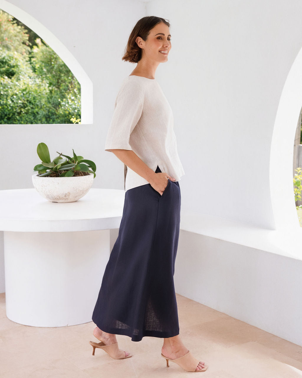 A woman modeling a natural linen short-sleeve top and navy blue wide-leg skirt in a bright, modern white room with arched windows and a small potted plant on a white table.