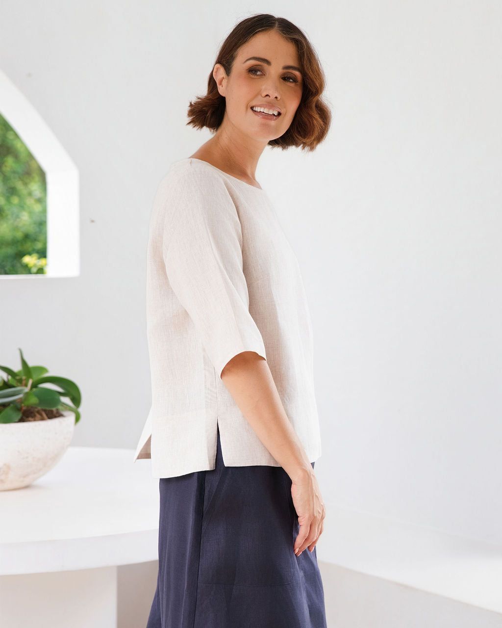 A woman modeling a natural linen See Saw three-quarter sleeve top and light white skirts , standing in a bright, modern white room with a round white table.