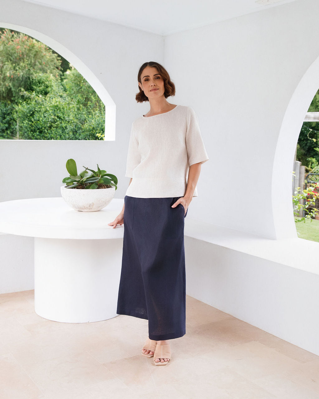 A woman modeling a natural linen See Saw three-quarter sleeve top and light white skirts , standing in a bright, modern white room with a round white table.