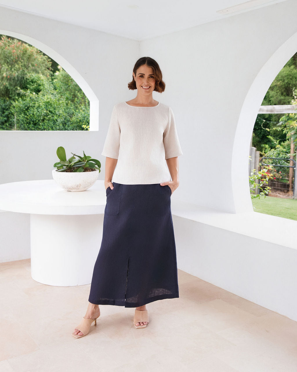 A woman modeling a cream short-sleeve knit top and a navy midi skirt, standing in a bright, modern white architectural space with arched windows and a round white table.