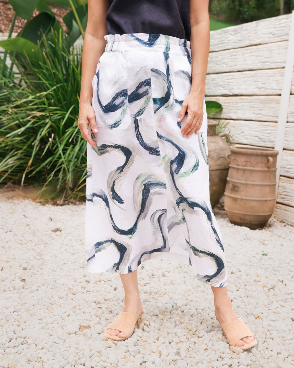A model wearing a white See Saw midi skirt with a dark navy blue riverstone abstract wavy print, paired with a black top and beige slide sandals, standing on a gravel path outdoors.