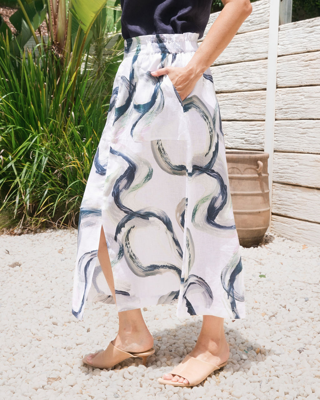 A model wearing a white See Saw midi skirt with a dark navy blue riverstone abstract wavy print, paired with a black top and beige slide sandals, standing on a gravel path outdoors.