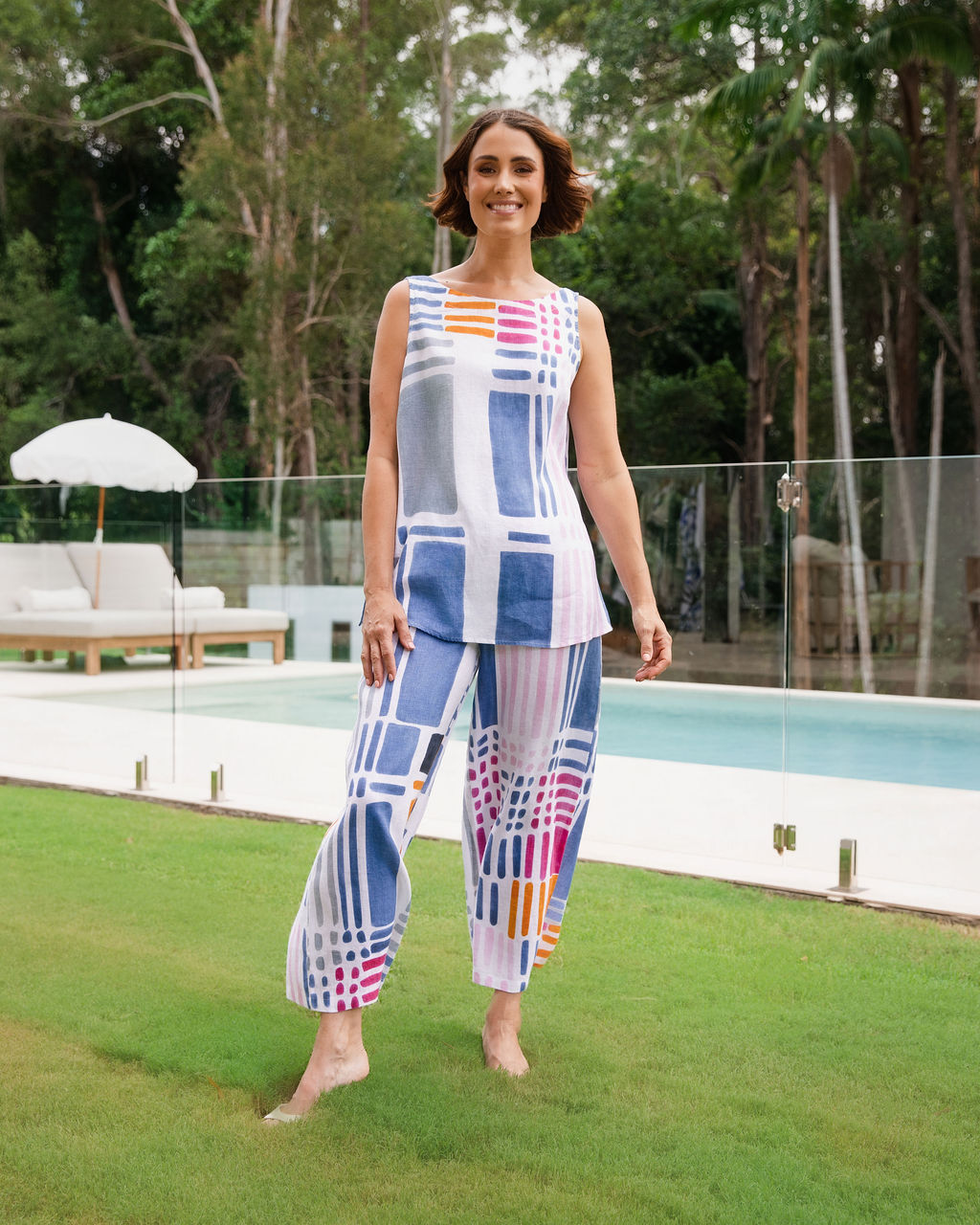 Woman modelling 100% Linen Gallery print boat neck tank by See Saw Clothing. The woman is standing outdoors in a pool area and garden setting. 