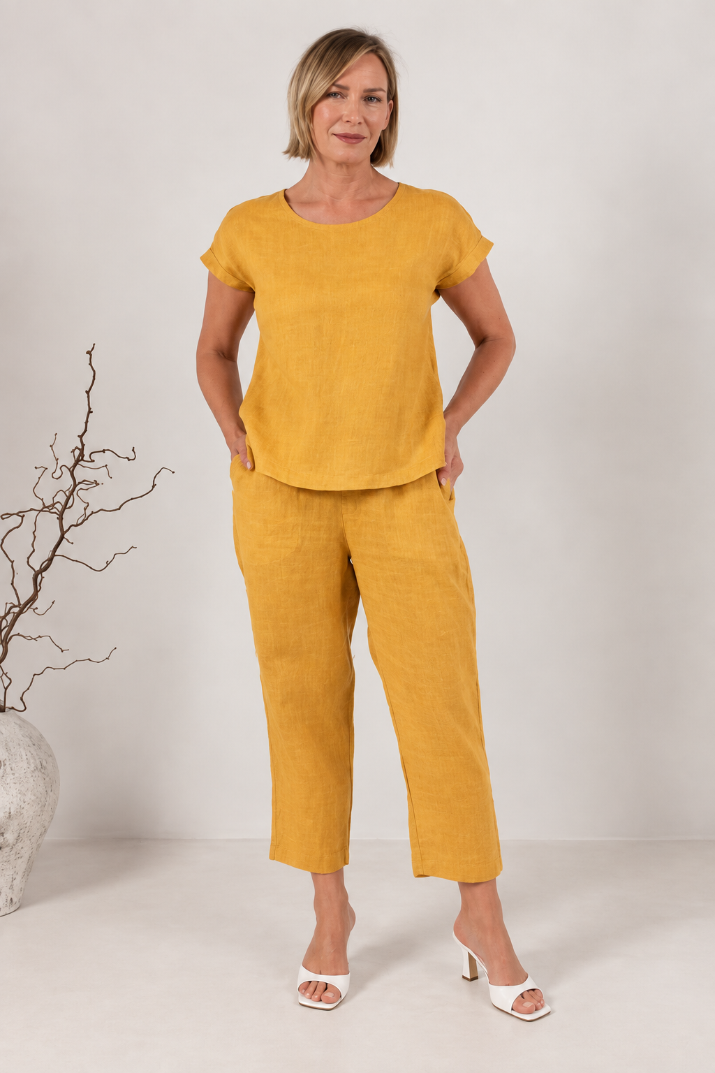 A smiling blonde woman wearing a marigold short-sleeve See Saw linen top and matching marigold pants, photographed against a plain light beige studio background.