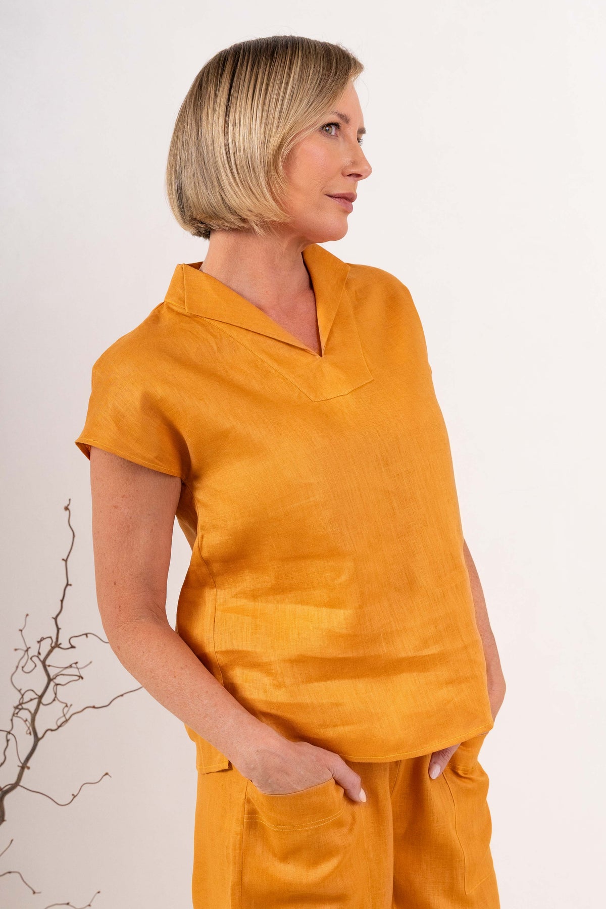 A woman modeling a marigold See Saw linen short-sleeve top with a split V-neck collar, paired with matching yellow linen pants, against a light off-white background.