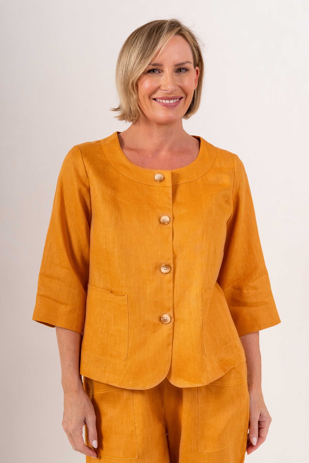 Woman modelling marigold linen see saw utility jacket top and white pants and against a plain background. 