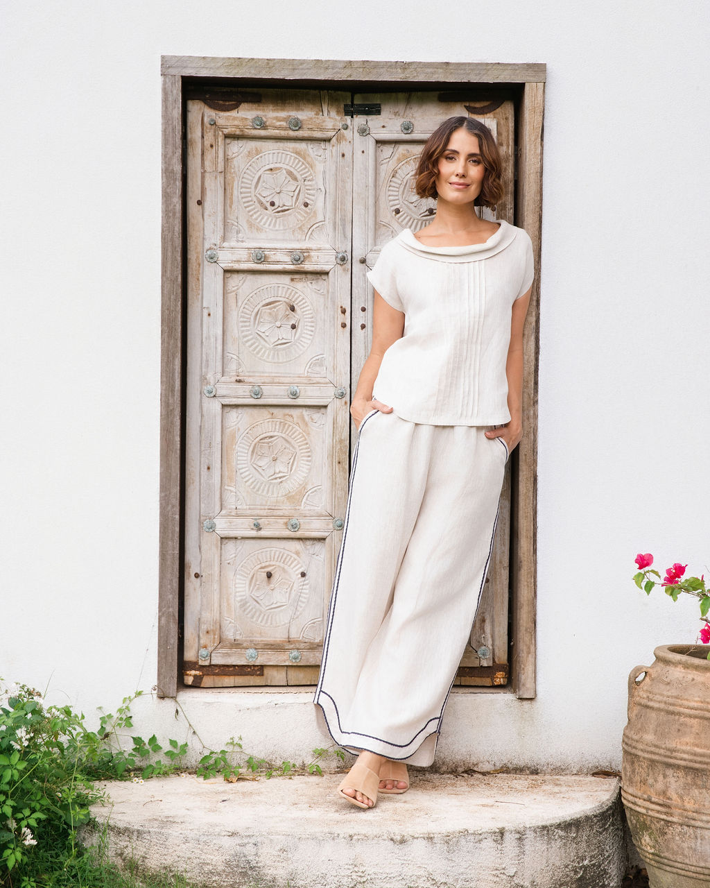 A woman stands barefoot on a stone step in front of a white wall and rustic wooden door, wearing a See Saw linen natural -colored short-sleeve knit top and matching wide-leg pants with dark piping.