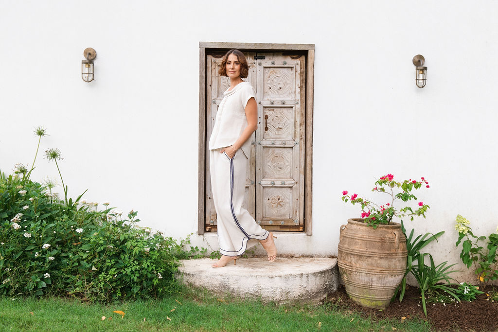 A woman stands barefoot on a stone step in front of a white wall and rustic wooden door, wearing a See Saw linen natural -colored short-sleeve knit top and matching wide-leg pants with dark piping.