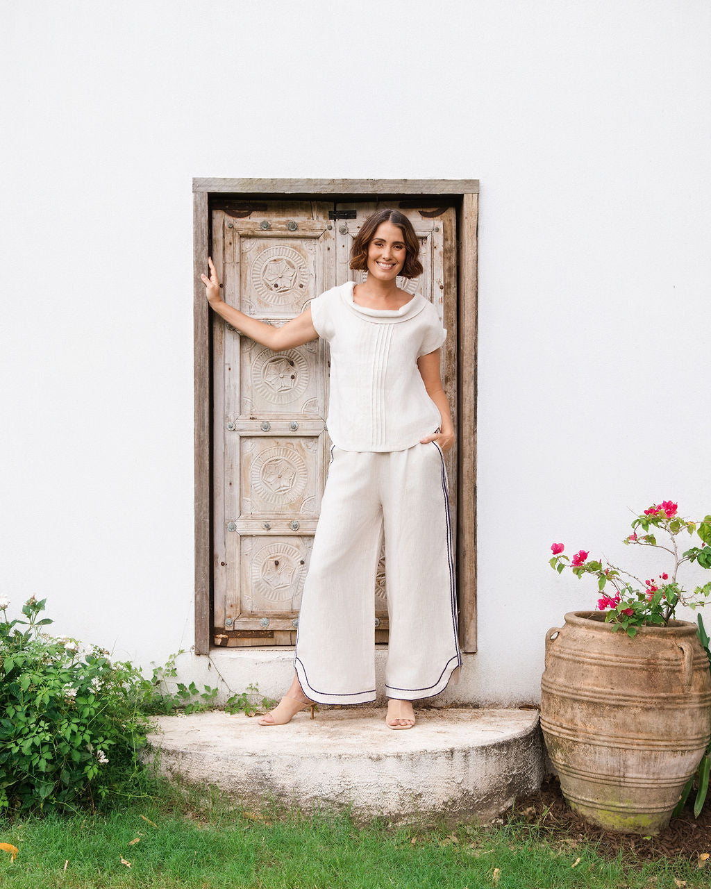 A woman stands barefoot on a stone step in front of a white wall and rustic wooden door, wearing a See Saw linen natural -colored short-sleeve knit top and matching wide-leg pants with dark piping.