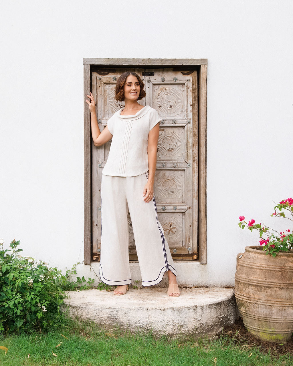 A woman stands barefoot on a stone step in front of a white wall and rustic wooden door, wearing a See Saw linen natural -colored short-sleeve knit top and matching wide-leg pants with dark piping.