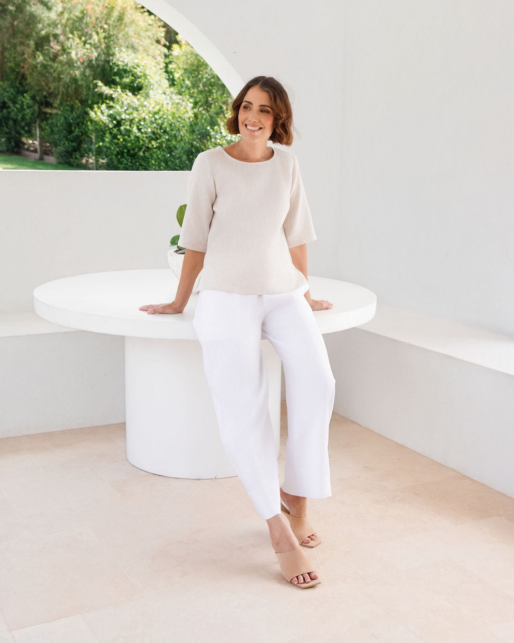 A woman modeling a natural linen See Saw three-quarter sleeve top and light white skirts , standing in a bright, modern white room with a round white table.