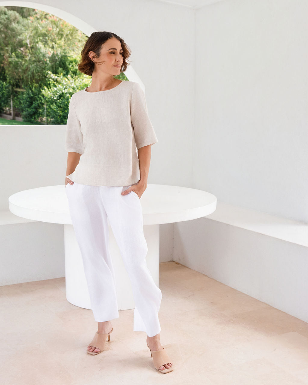 A woman modeling a natural linen See Saw three-quarter sleeve top and light white skirts , standing in a bright, modern white room with a round white table.