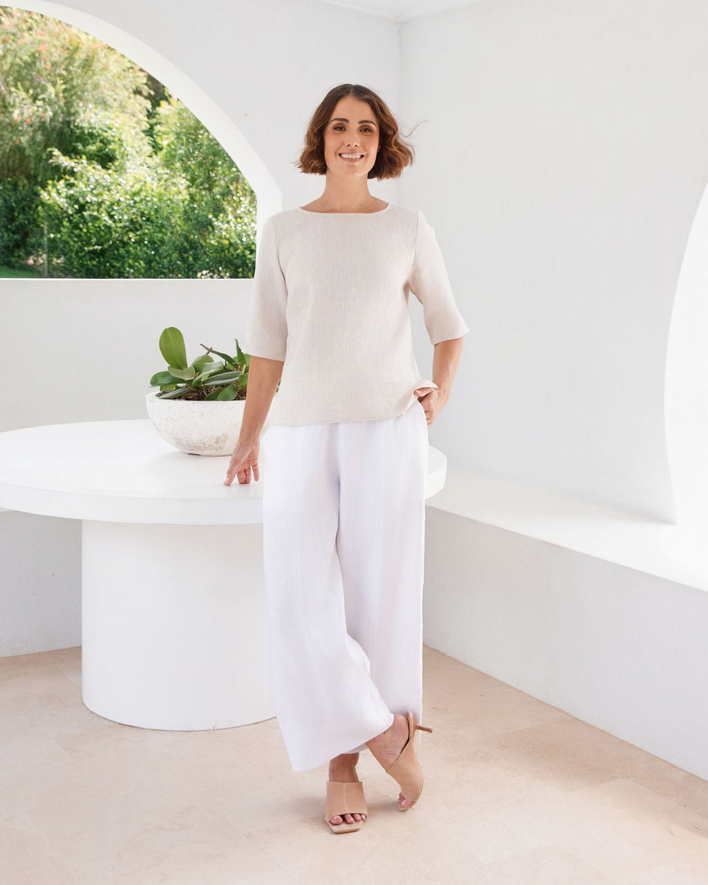 A woman modeling a natural linen See Saw three-quarter sleeve top and light white skirts , standing in a bright, modern white room with a round white table.