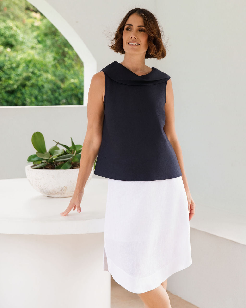 A woman modeling a sleeveless linen See Saw dark navy top and white skirt standing in a bright, modern white room with a curved table and greenery visible through an arched window.