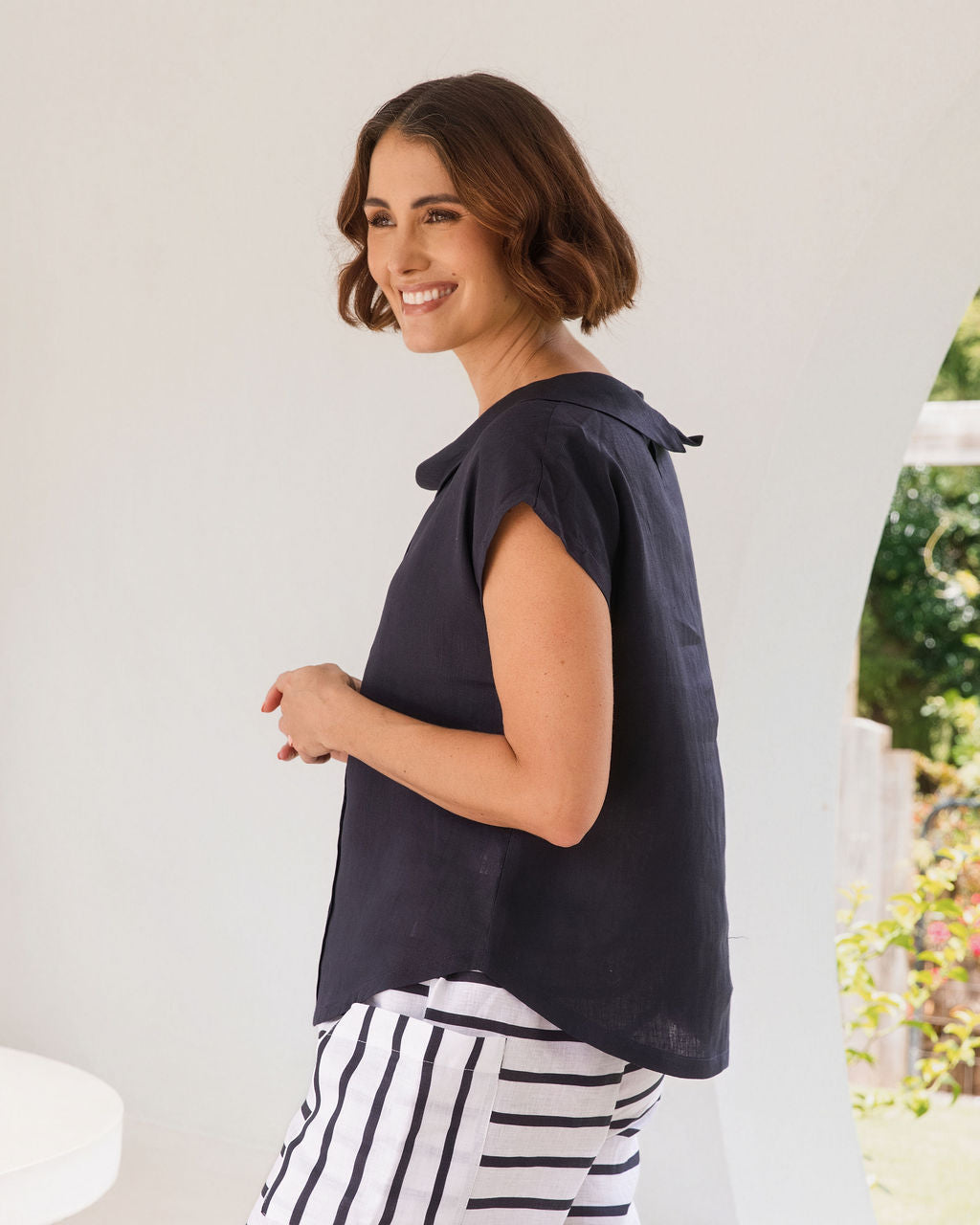 A woman stands in a bright, modern white architectural space with arched windows overlooking greenery, wearing a dark navy linen See Saw top sleeveless top and navy-and-white horizontally striped cropped pants.