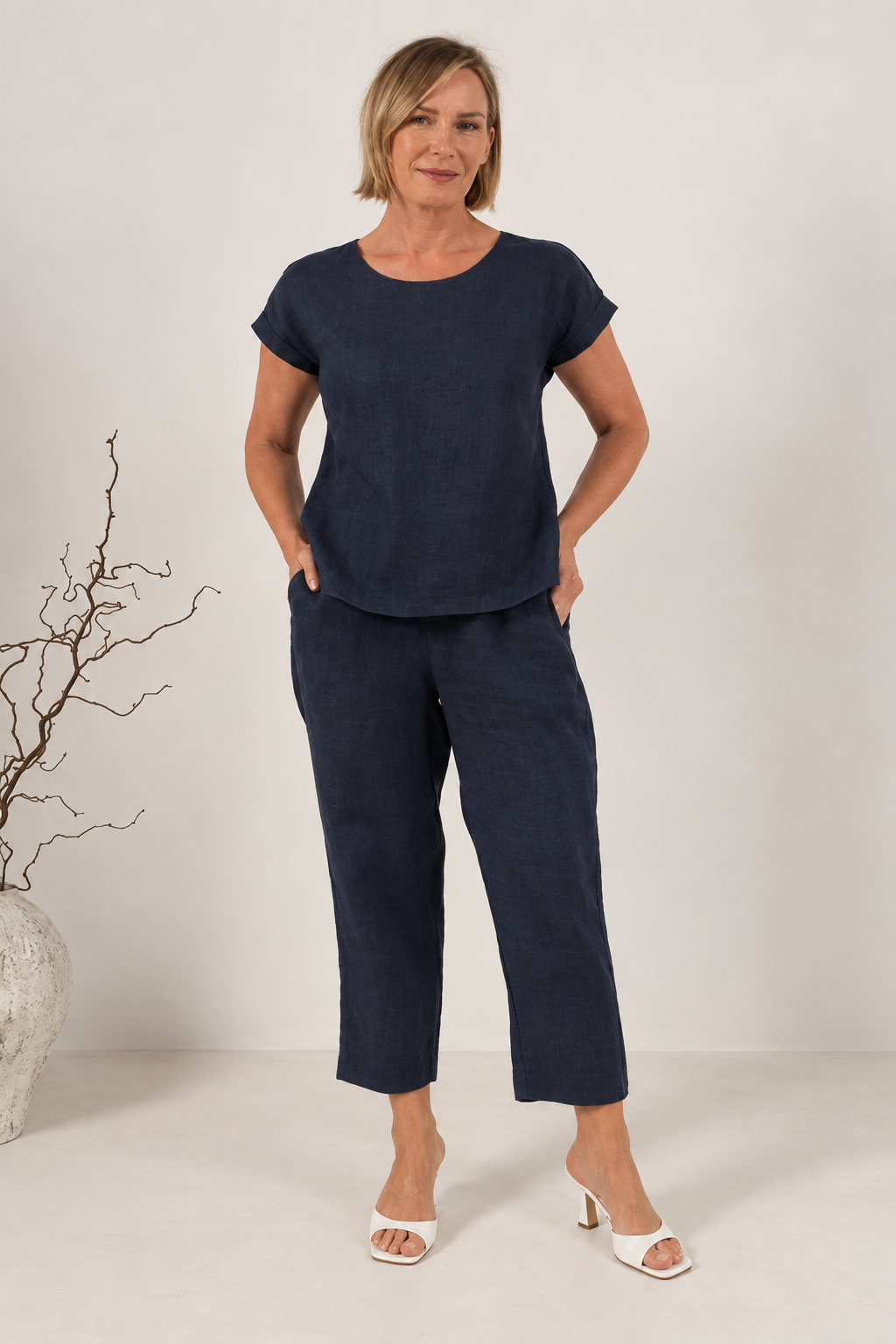 A smiling blonde woman wearing a navy short-sleeve See Saw linen top and matching navy pants, photographed against a plain light beige studio background.