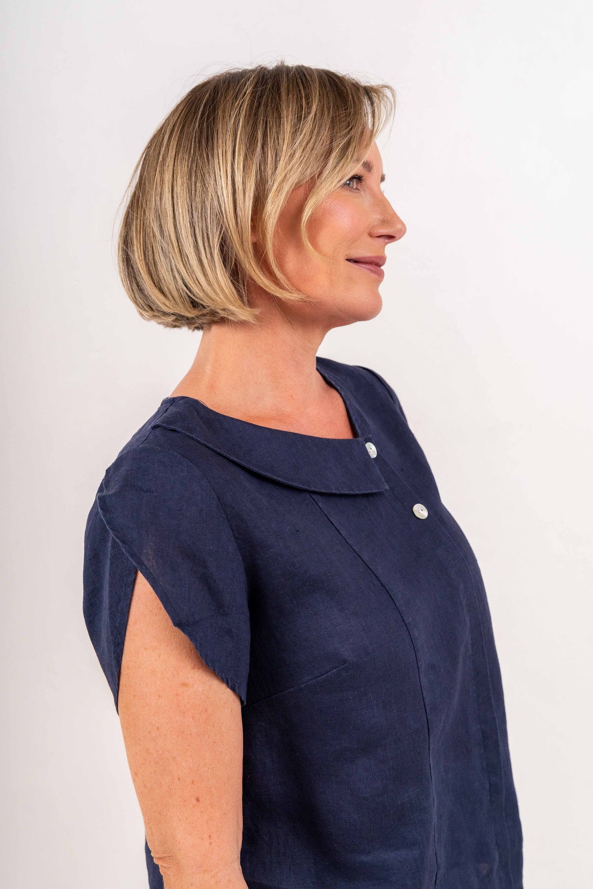 A woman with a short blonde bob is shown in profile wearing a navy blue See Saw linen short-sleeve top with a collar and silver buttons, photographed against a plain light background.