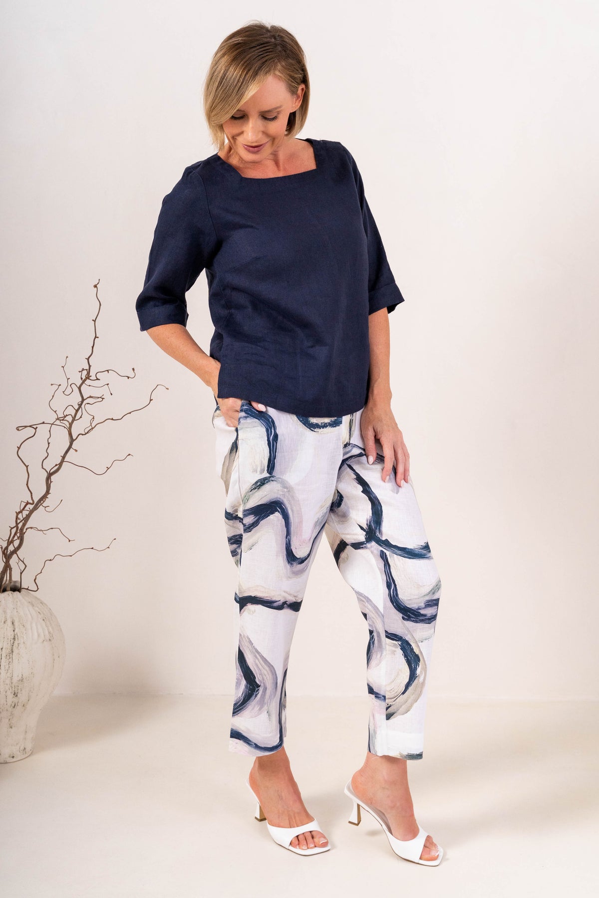 A woman wearing a navy linen See Saw top with a square neckline and elbow-length sleeves, paired with colorful riverstone pants, standing in a studio.