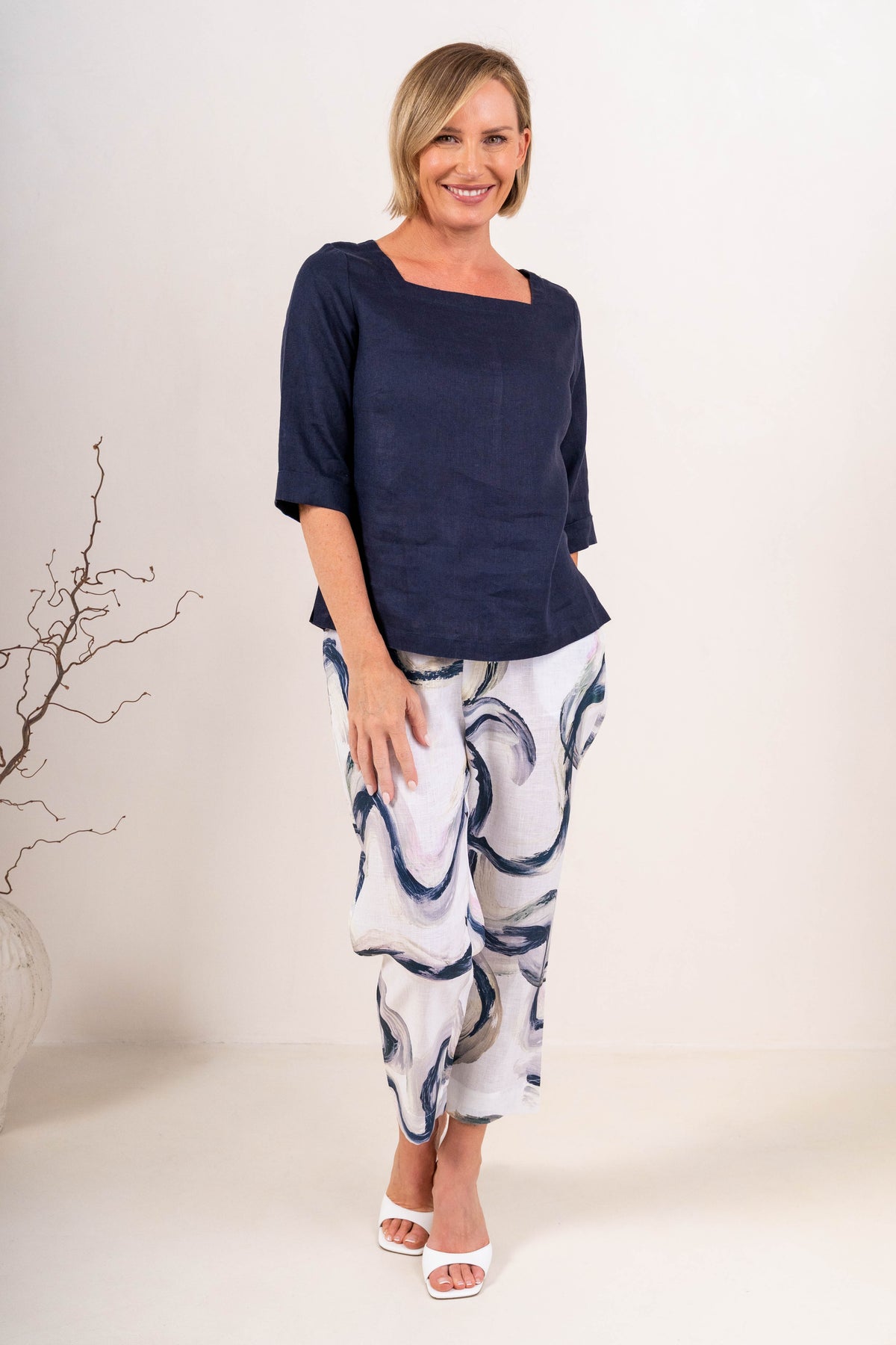 A woman wearing a navy linen See Saw top with a square neckline and elbow-length sleeves, paired with colorful riverstone pants, standing in a studio.