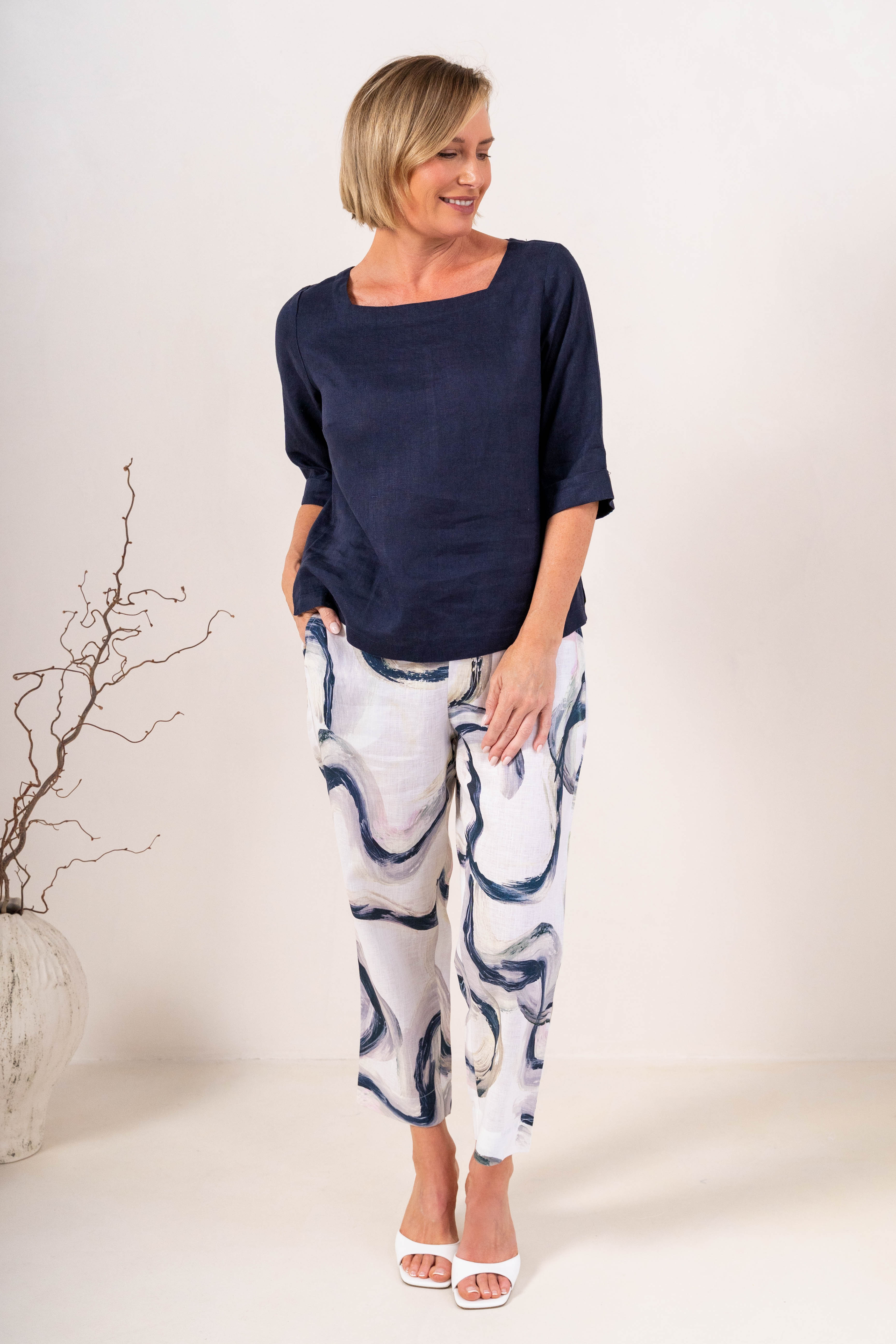 A woman wearing a navy linen See Saw top with a square neckline and elbow-length sleeves, paired with colorful riverstone pants, standing in a studio.