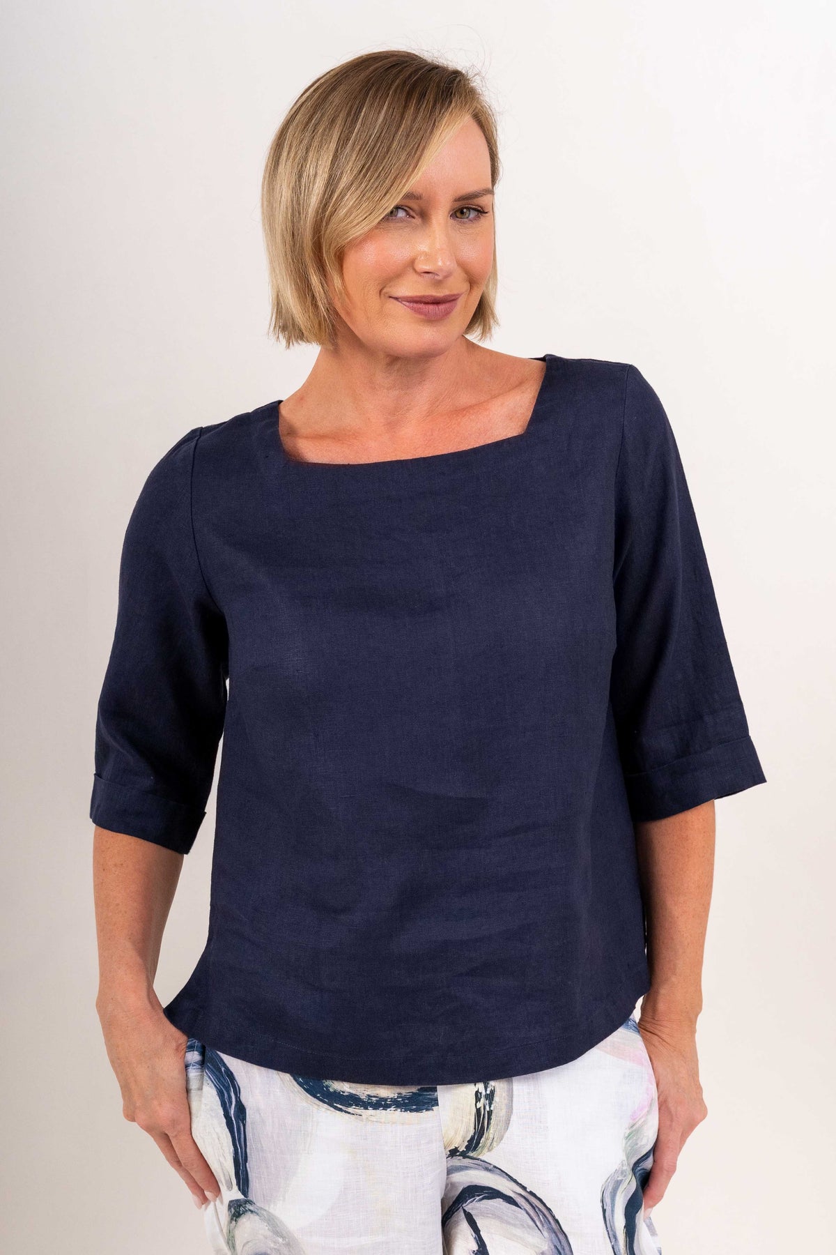 A woman wearing a navy linen See Saw top with a square neckline and elbow-length sleeves, paired with colorful riverstone pants, standing in a studio.