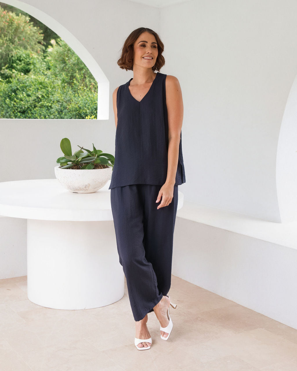 Model wearing See Saw Matching tank and pant in navy in linen and rayon blend. standing in an outdoor architecturally designed patio.