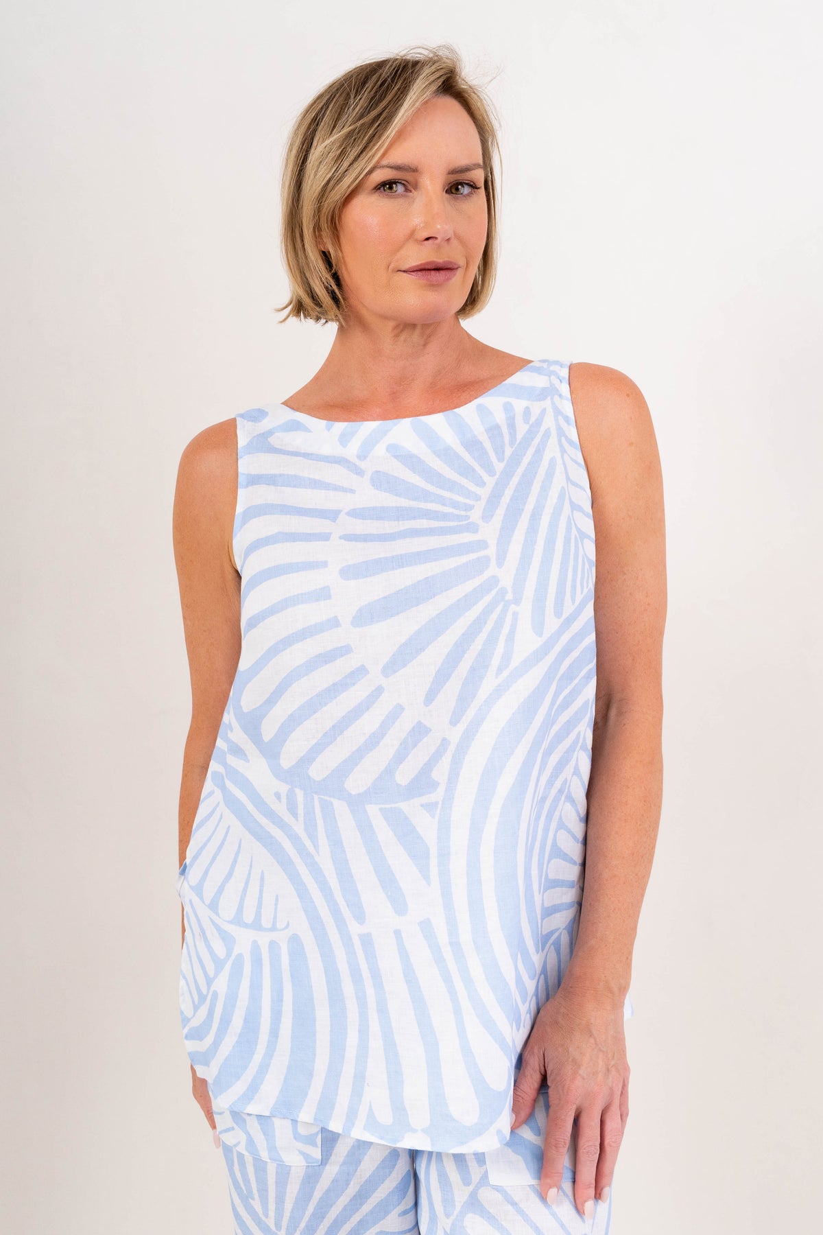 A woman modeling a sleeveless light Palm breeze print See Saw linen patterned top against a plain light background.