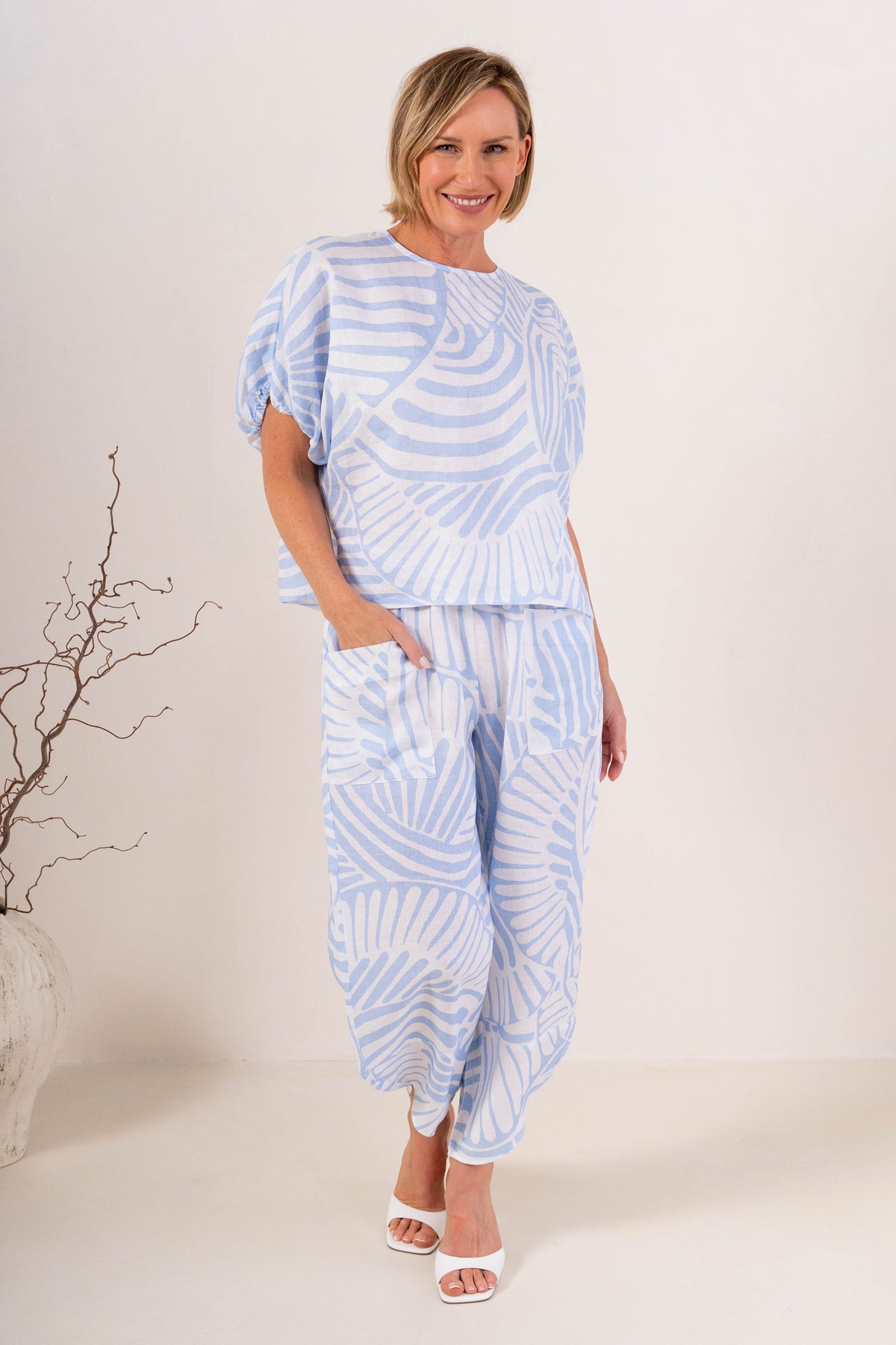 A woman modeling a Palm Breeze linen patterned two-piece set consisting of a short-sleeve top and matching pants, standing against a plain off-white studio background.