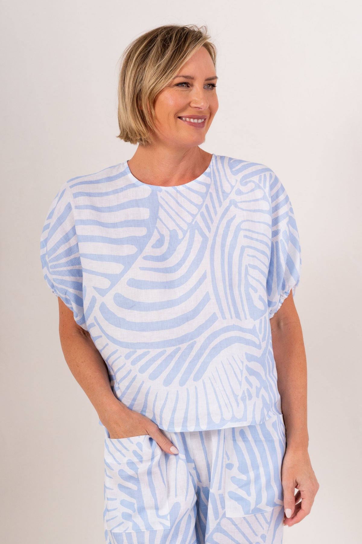 A woman modeling a Palm Breeze linen patterned two-piece set consisting of a short-sleeve top and matching pants, standing against a plain off-white studio background.