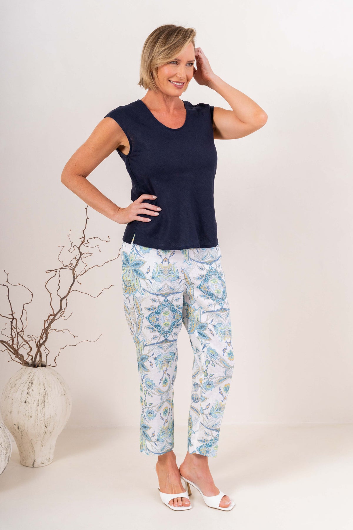 A smiling blonde woman wearing a navy sleeveless V-neck top in linen by See Saw and matching white pants, standing against a plain light background.
