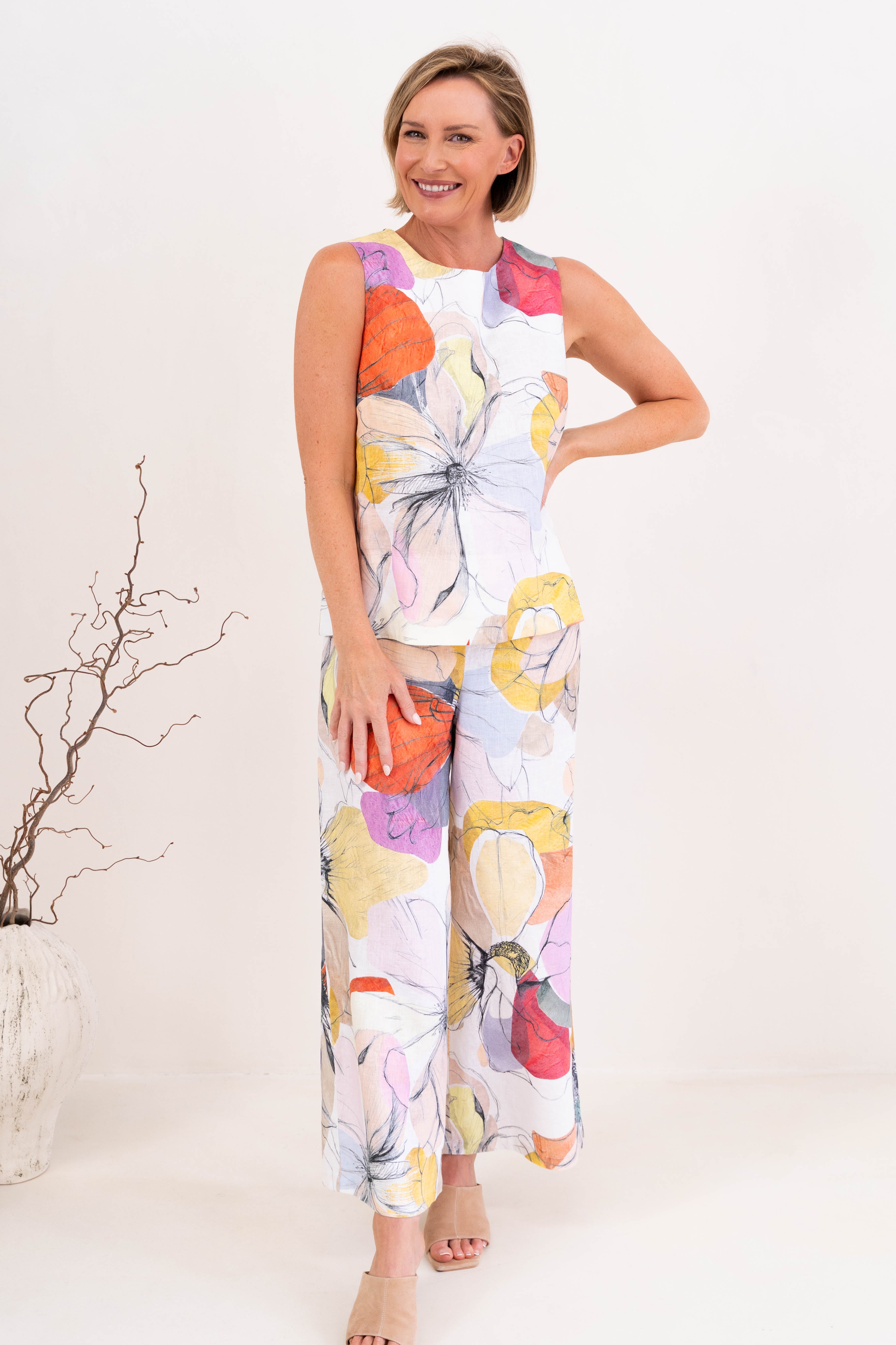 A woman modeling a sleeveless Petal Drift linen See Saw Top and pant set with a colorful abstract floral print, standing in a bright, modern living room.