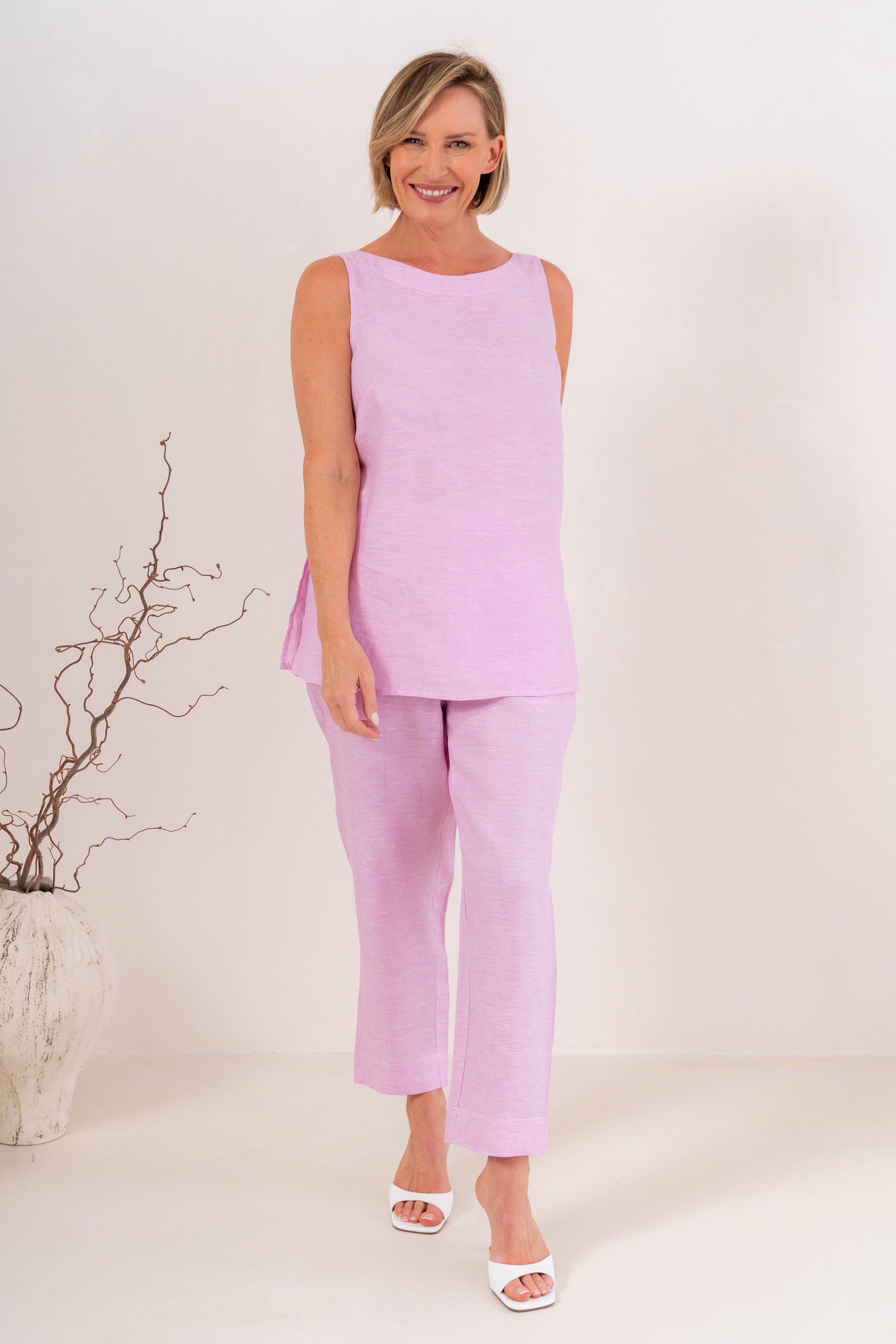 A person wearing light pink linen  See Saw 7/8 pants with a relaxed straight-leg fit, paired with white heeled slide sandals, photographed against a plain light beige studio background.