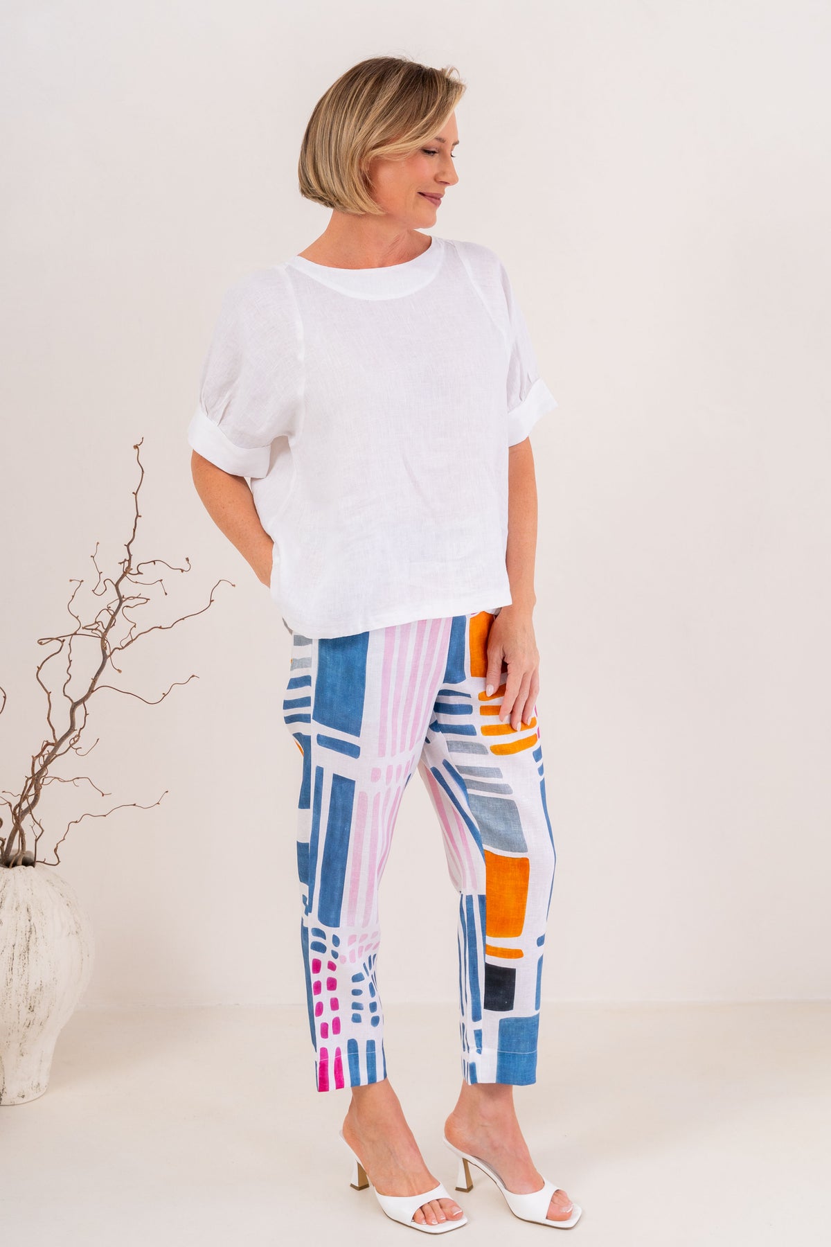 A woman modeling a white linen See Saw short-sleeve top and colorful geometric print cropped pants, standing against a plain off-white studio background with a decorative vase of branches on the left.