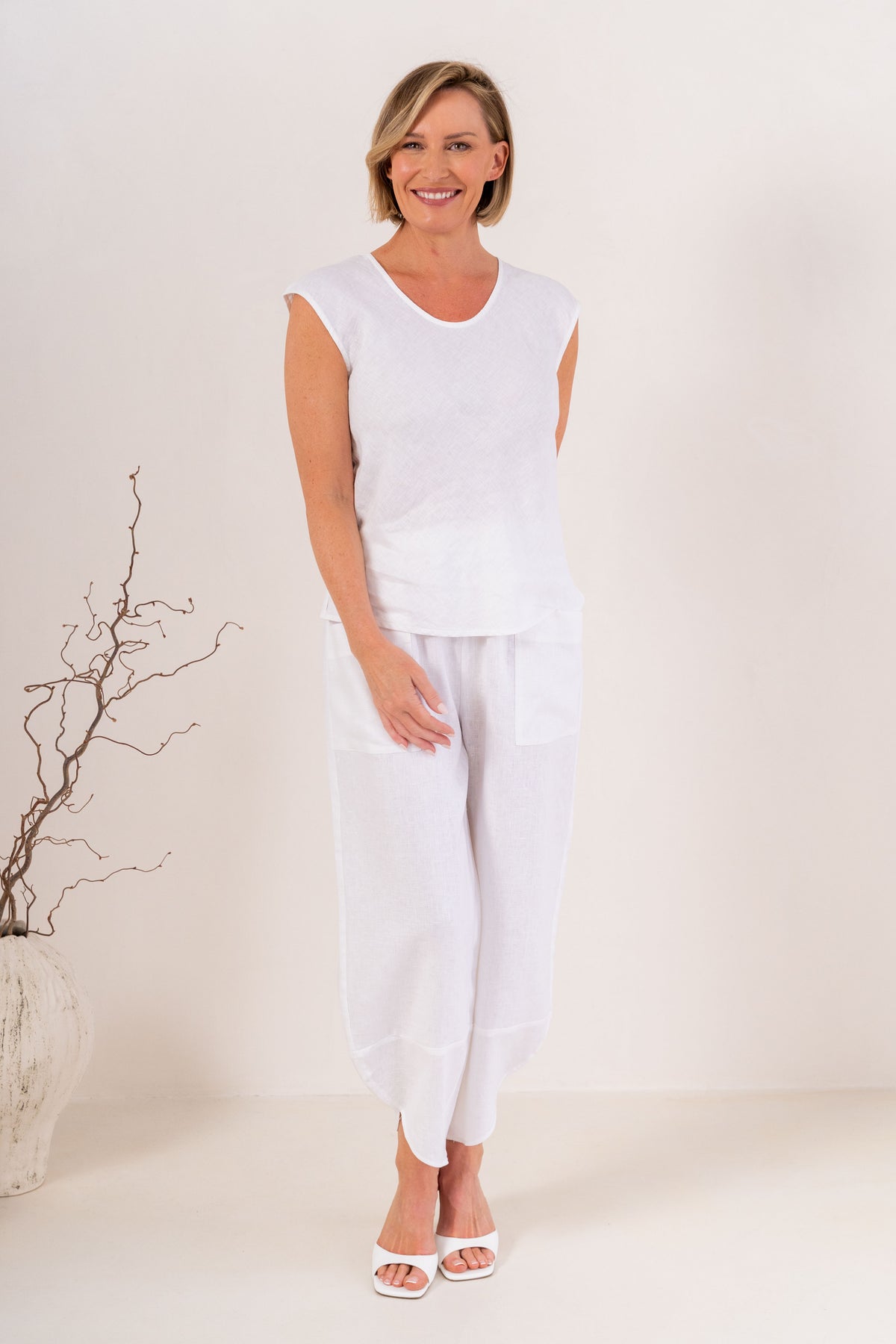 A smiling blonde woman wearing a white sleeveless V-neck top in linen by See Saw and matching white pants, standing against a plain light background.