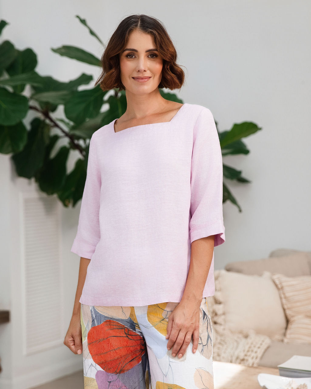A woman wearing a light pink linen See Saw top with a square neckline and elbow-length sleeves, paired with colorful petal drift pants, standing in a bright living room.