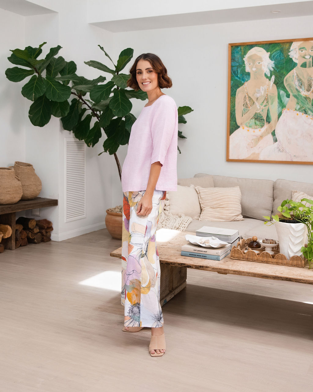 A woman wearing a light pink linen See Saw top with a square neckline and elbow-length sleeves, paired with colorful petal drift pants, standing in a bright living room.