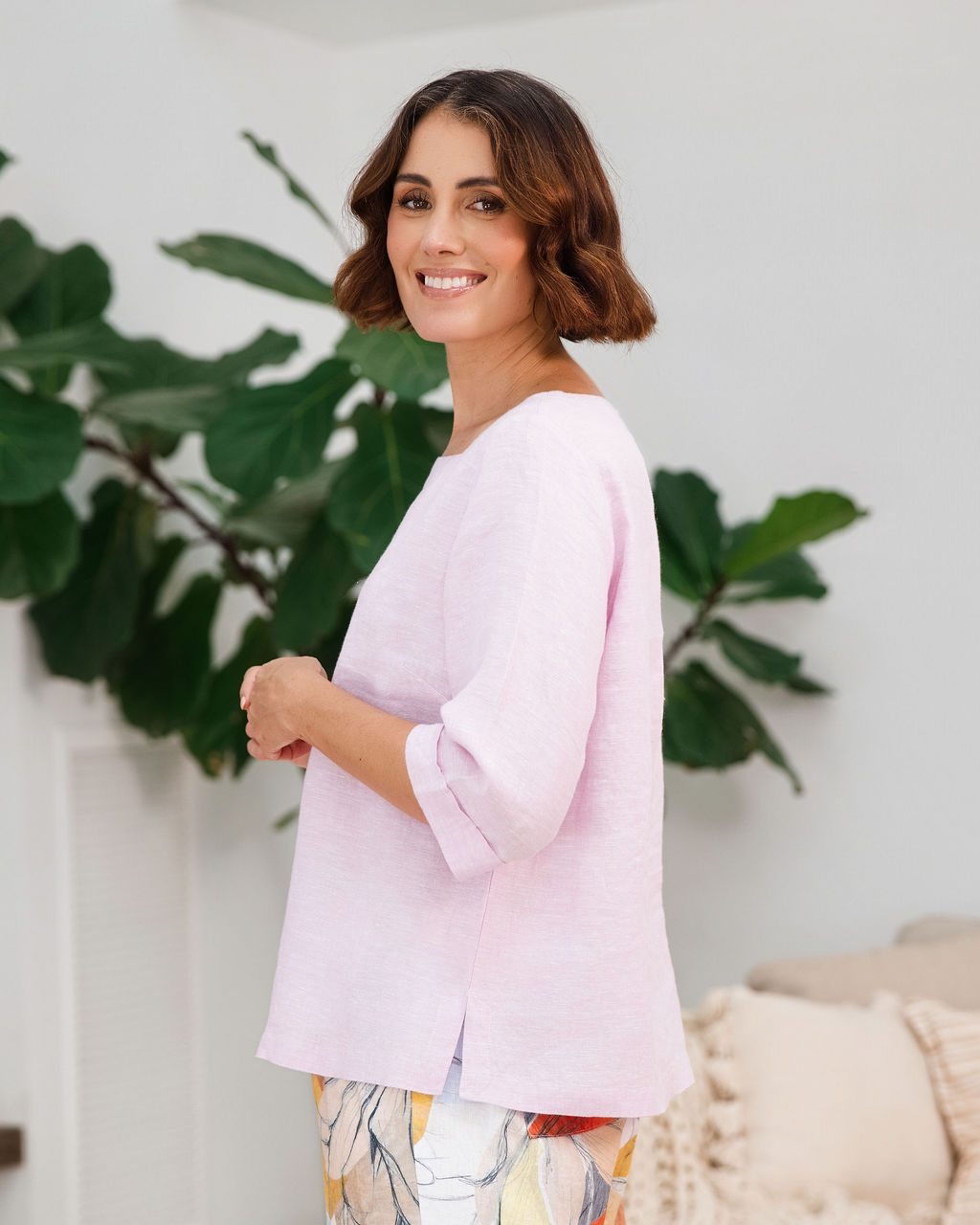 A woman wearing a light pink linen See Saw top with a square neckline and elbow-length sleeves, paired with colorful petal drift pants, standing in a bright living room.